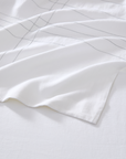 A close-up of a white fabric textile, likely a bed sheet or duvet cover, featuring a minimalist thin gray grid pattern, draped elegantly on a matching white background.