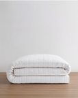 A folded white quilted comforter or duvet with a smooth, padded texture, stacked neatly on a light brown wooden surface against a plain off-white wall.