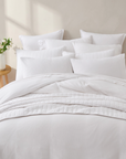 A neatly made bed with white quilted bedding, including a duvet cover, multiple white pillows, and a matching pillowcase, styled in a bright, minimalist bedroom setting with a small wooden side table and flowers.