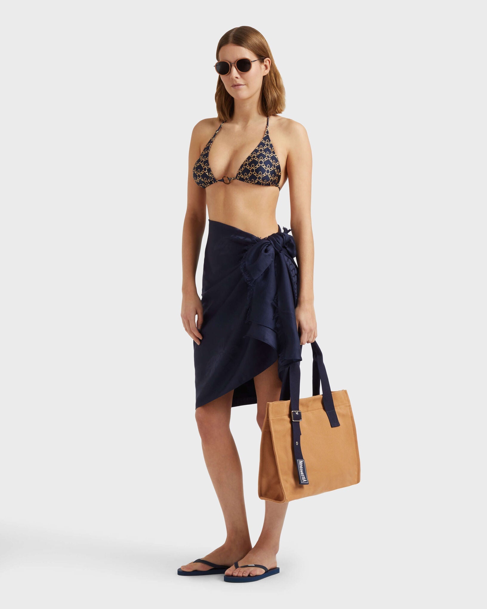 Model wearing the Vilebrequin Turtles Chain bikini top styled with a navy sarong, sunglasses, and a tan canvas beach tote bag.