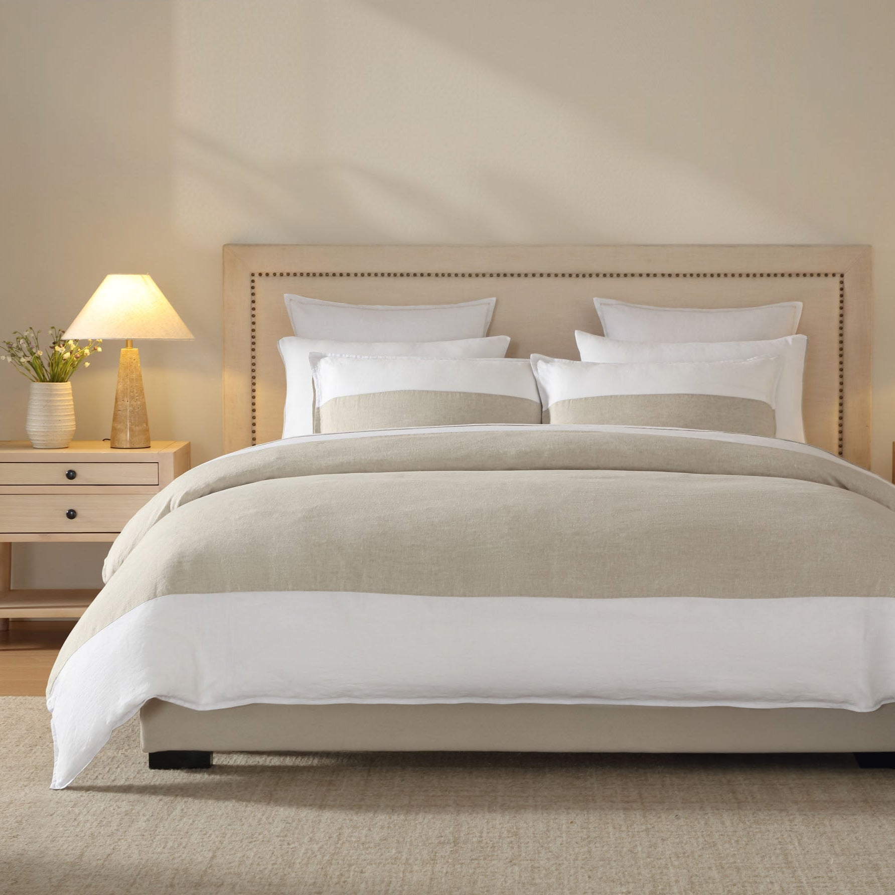 A bed with a beige linen quilt cover, flanked by two nightstands with lamps, in a room with a beige wall and wooden flooring.