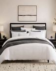 A neatly made bed in a modern bedroom setting, featuring a white quilted duvet cover, white pillows, and a dark gray textured throw blanket. The bed is supported by a black metal frame with a headboard, flanked by matching black nightstands with lit table lamps.