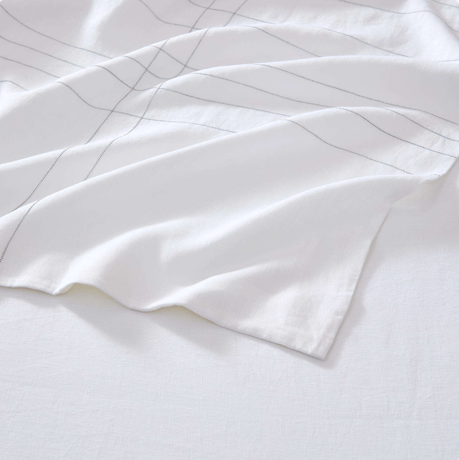 A close-up of a white fabric textile, likely a bed sheet or duvet cover, featuring a minimalist thin gray grid pattern, draped elegantly on a matching white background.