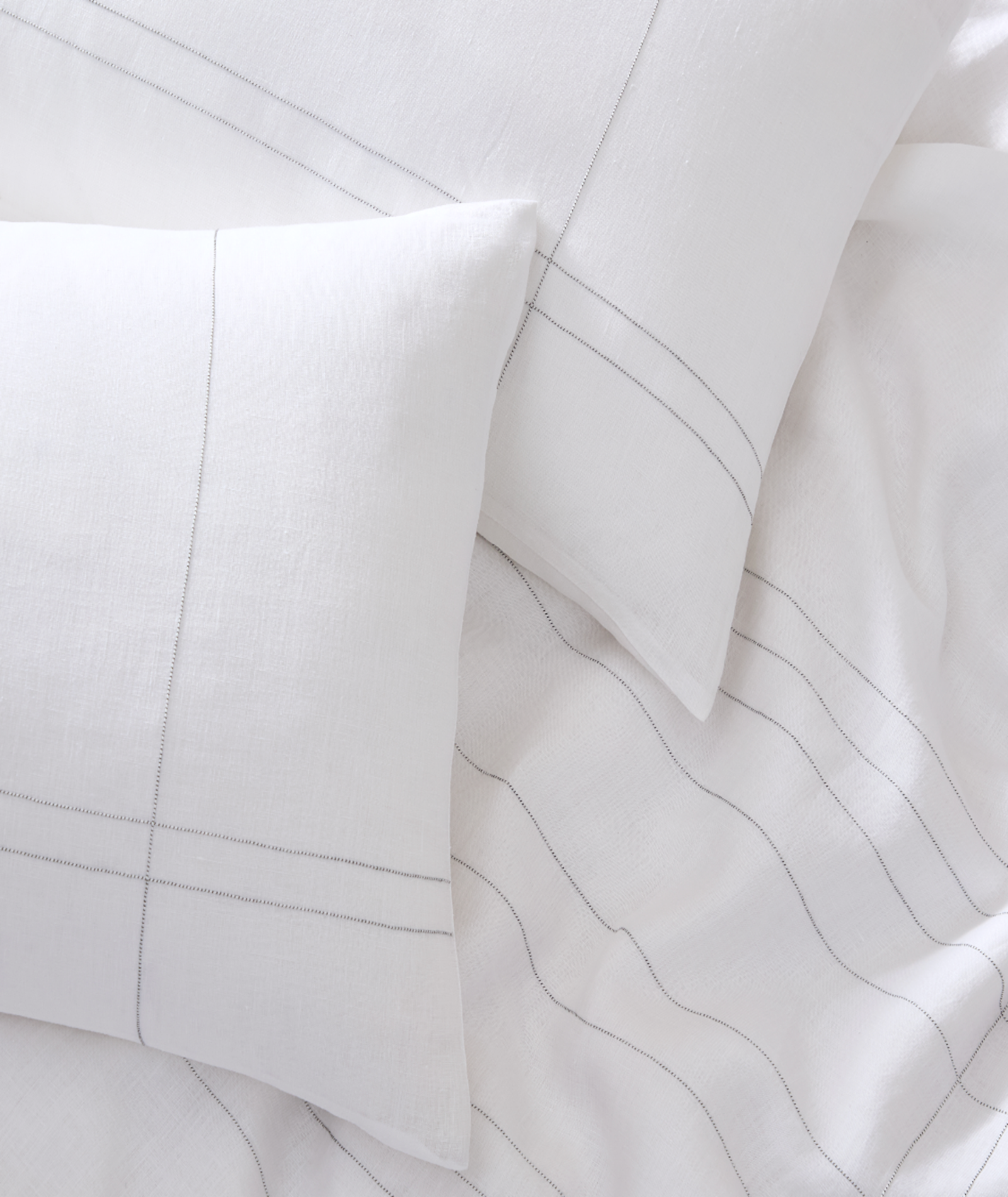 White cotton bedding set with two pillows and a duvet cover featuring delicate thin gray stitched lines, arranged in a styled flat-lay composition.