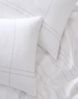 White cotton bedding set with two pillows and a duvet cover featuring delicate thin gray stitched lines, arranged in a styled flat-lay composition.