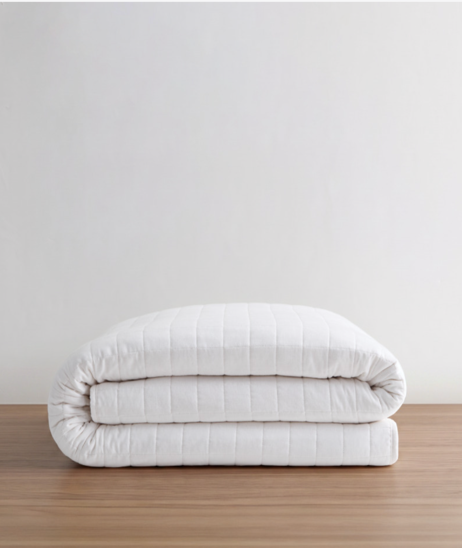 A folded white quilted comforter or duvet with a smooth, padded texture, stacked neatly on a light brown wooden surface against a plain off-white wall.
