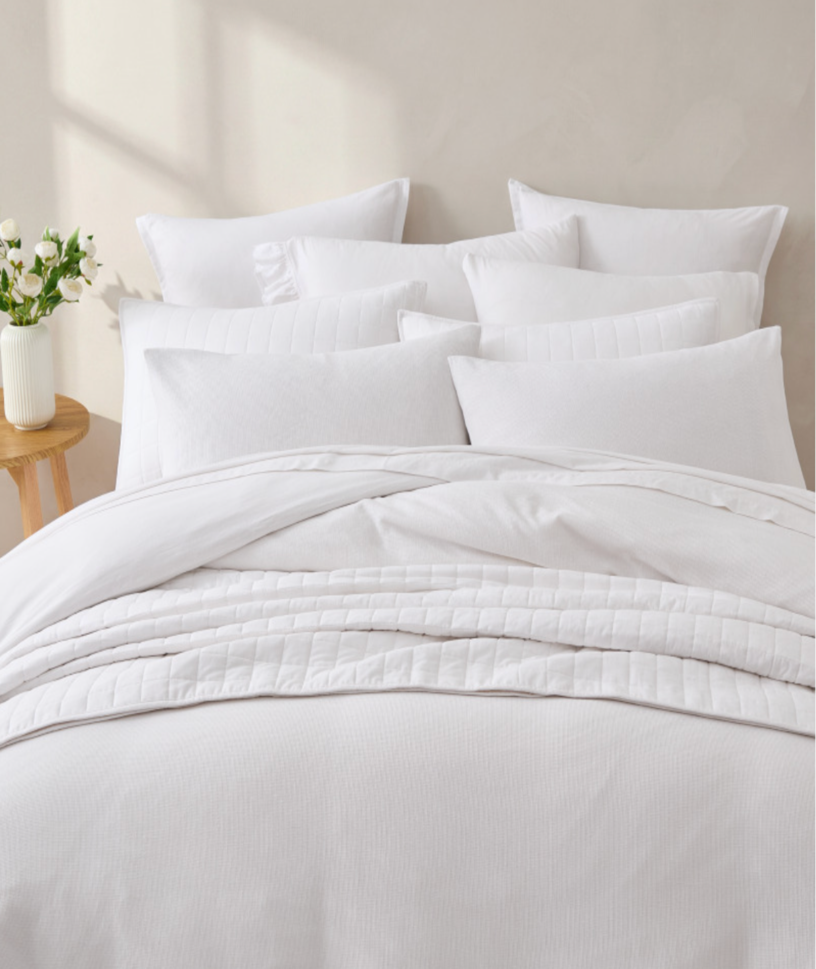 A neatly made bed with white quilted bedding, including a duvet cover, multiple white pillows, and a matching pillowcase, styled in a bright, minimalist bedroom setting with a small wooden side table and flowers.